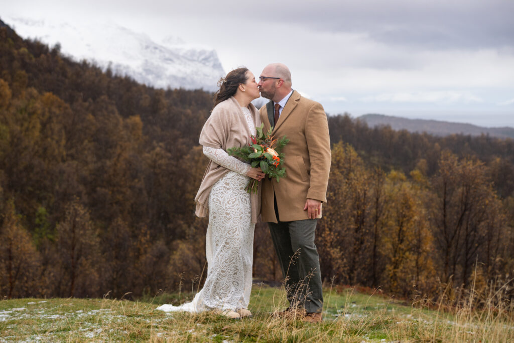 Intimate Norway adventure elopement moment as couple kisses on a mountainside with snow-capped peaks, golden autumn forest, and moody skies — captured by Tromsø adventure elopement photographer