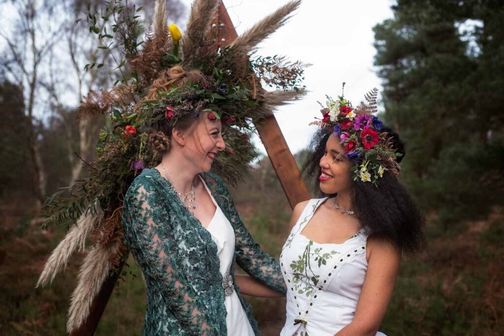 So, should we elope? Deciding to elope is a deeply personal choice for any couple. This image captures the essence of an intimate, romantic elopement with two brides smiling with excitement at each other on their elopement day amidst a natural, serene forest setting. Their flower-adorned hairstyles and unique wedding attire reflect the personalized, meaningful experience that an elopement can offer. If the idea of a cozy, personal ceremony away from the hustle and bustle of a large wedding resonates with you, eloping might be the perfect choice.